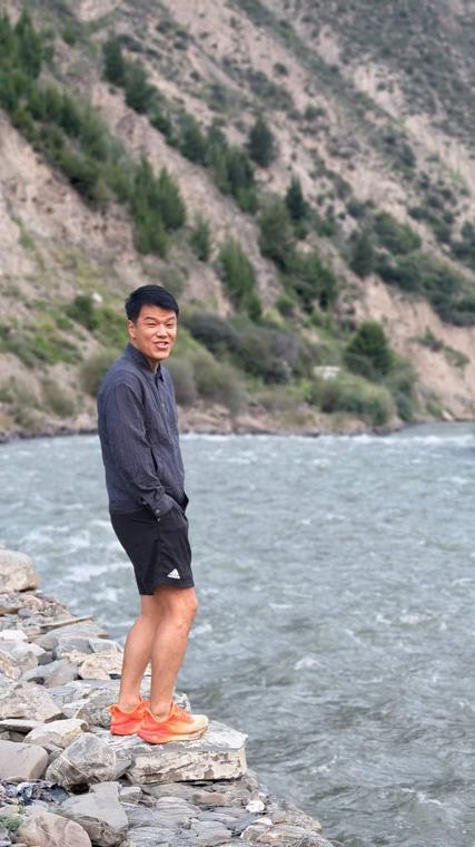 朱老师站在河边的石头上，身后是山坡