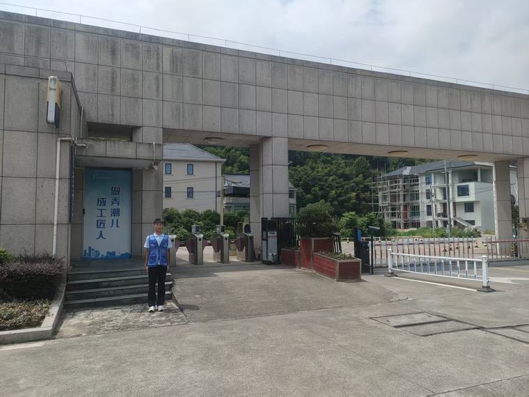 学校大门前，一位穿蓝色马甲的同学站在远处，画面中显得很小