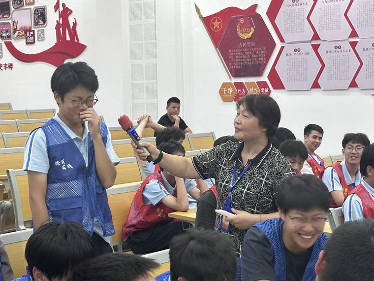 活动现场，老师拿着话筒采访一位站起来的同学，周围同学在笑