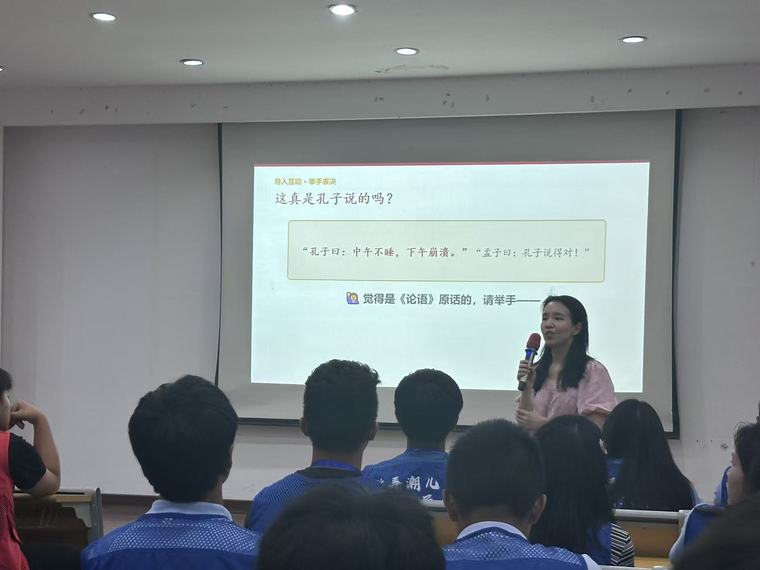 老师拿着话筒站在投影屏幕旁讲课，同学们坐在台下